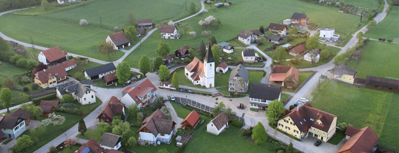 Luftaufnahme von Stiwoll mit verstreuten Häusern, grünen Feldern und einer weißen Kirche mit einem hohen Kirchturm in der Mitte, umgeben von Straßen und Bäumen.