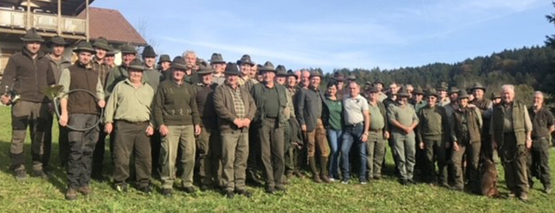 Eine große Gruppe von Menschen, meist Männer, steht im Freien auf einer Wiese vor einem Gebäude und Bäumen. Viele sind mit grüner oder brauner Outdoor-Kleidung und Hüten bekleidet, und auf der rechten Seite der Gruppe ist ein Hund zu sehen.