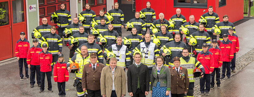 Eine große Gruppe von Feuerwehrmännern in gelb gestreiften Uniformen und Helmen steht in Reihen vor einer Feuerwache. Mehrere Personen in förmlicher Kleidung und roten Jacken stehen an der Spitze.