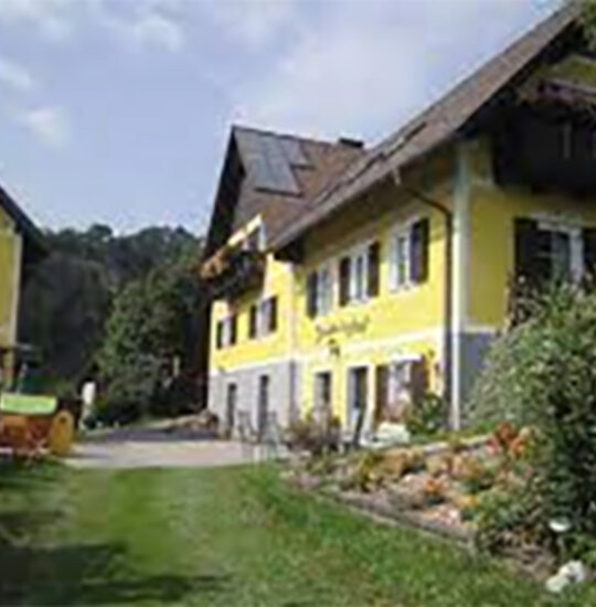 Ein gelbes Haus mit einem dunklen Dach steht auf einem grasbewachsenen Hügel, umgeben von Bäumen und Bergen unter einem teilweise bewölkten Himmel. Die Landschaft ist üppig und grün, und im Hintergrund sind ferne Hügel zu sehen.