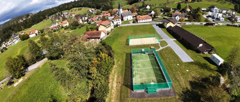 Luftaufnahme eines kleinen ländlichen Dorfes mit Häusern, grünen Feldern und einem eingezäunten Sportplatz, umgeben von Bäumen und Hügeln unter einem teilweise bewölkten Himmel.