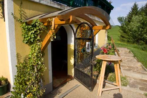 Eine rustikale Eingangstür mit einem dekorativen Eisentor, einer Holzmarkise und Kletterpflanzen. Draußen steht ein runder Holztisch neben einem Steinweg, der durch einen grünen Hof führt.