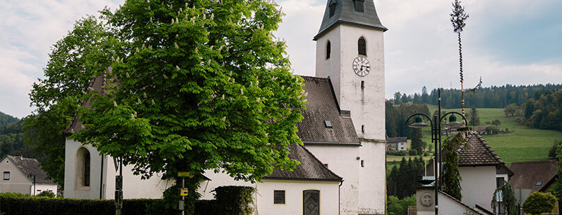 Eine weiße Kirche mit einem hohen Glockenturm und einem dunklen Kirchturm steht an einer Straße.
