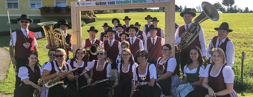Eine große Gruppe von Männern und Frauen in traditioneller bayerischer Kleidung posiert im Freien mit Musikinstrumenten und lächelt unter einem Holzschild inmitten sonniger grüner Felder.