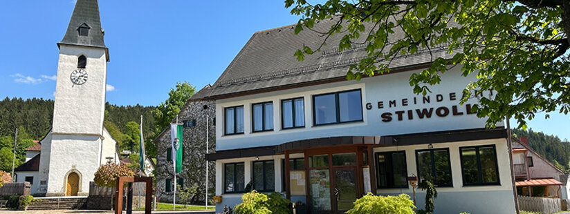 Ein weißes Gebäude mit der Aufschrift Gemeinde Stiwoll und großen Fenstern steht unter einem Baum, daneben eine weiße Kirche mit einem Glockenturm, vor einem Hintergrund aus Bäumen und blauem Himmel.