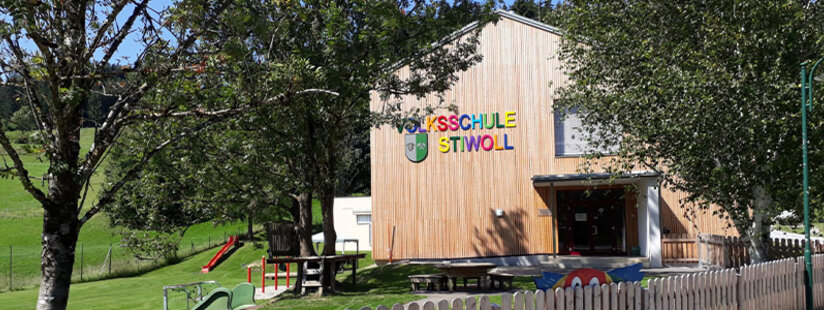 Ein modernes, zweistöckiges Schulgebäude aus Holz mit bunten Fensterrahmen und der Aufschrift Volksschule Stiwoll an der Fassade. Bäume und Grünflächen umgeben das Gebäude.