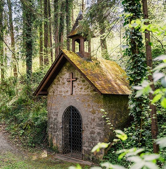 Eine kleine Steinkapelle mit einem Holzdach und einem Kreuz über dem gewölbten Eingang steht in einem üppigen, grünen Wald, umgeben von hohen Bäumen und dichtem Laub.