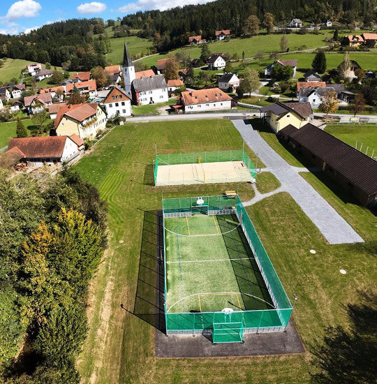 Luftaufnahme von Stiwoll mit grünen Feldern, Häusern, einer Kirche und zwei eingezäunten Sportplätzen - einem für Fußball und einem für Volleyball - zwischen Bäumen und Straßen an einem sonnigen Tag.