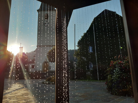 Regentropfen auf einem Fenster mit Blick auf einen Kirchturm, eine Uhr und ein nahe gelegenes Gebäude, im Hintergrund ist Sonnenlicht zu sehen.